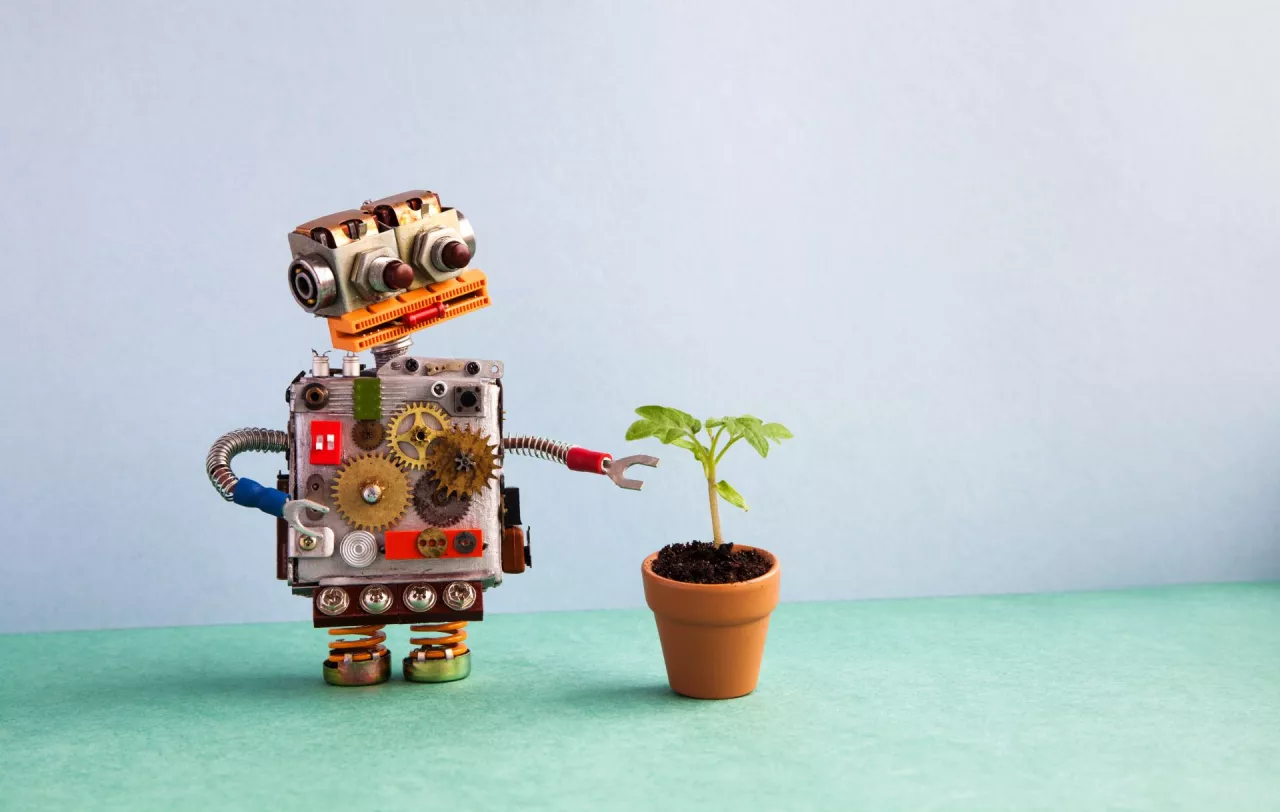 Robot and flowerpot. Creative design robotic character looks at the green plant housepot. Gray wall green floor background, copy space.